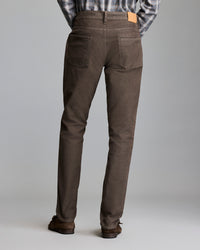 A person wearing brown pants, brown shoes, and a plaid shirt stands facing away from the camera against a plain gray background.