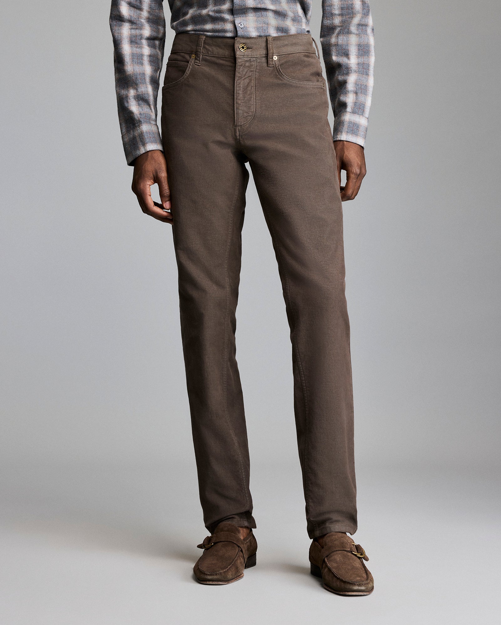 A person wearing brown straight-leg pants, a grey and white plaid shirt, and brown suede loafers stands against a plain light grey background. Only the lower half of the body is visible.