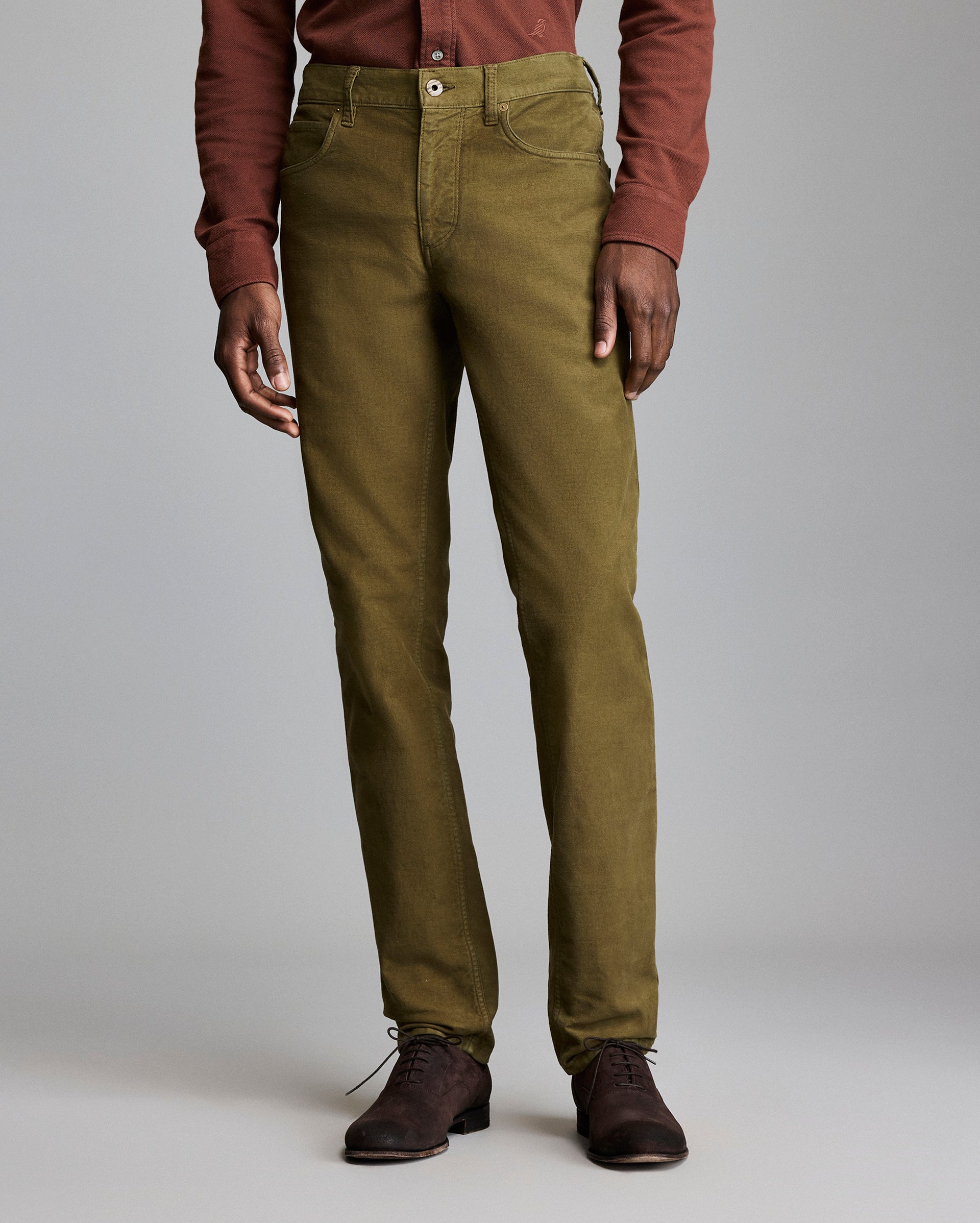 A person wearing olive green straight-leg pants, a rust-colored long-sleeve shirt, and dark brown shoes stands against a plain gray background.