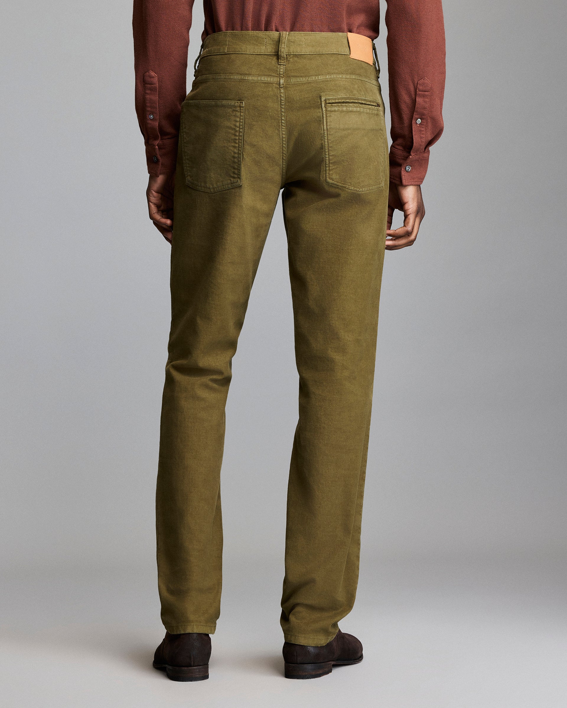 A person stands facing away, wearing olive green pants, a brown long-sleeve shirt, and dark brown shoes against a plain gray background.
