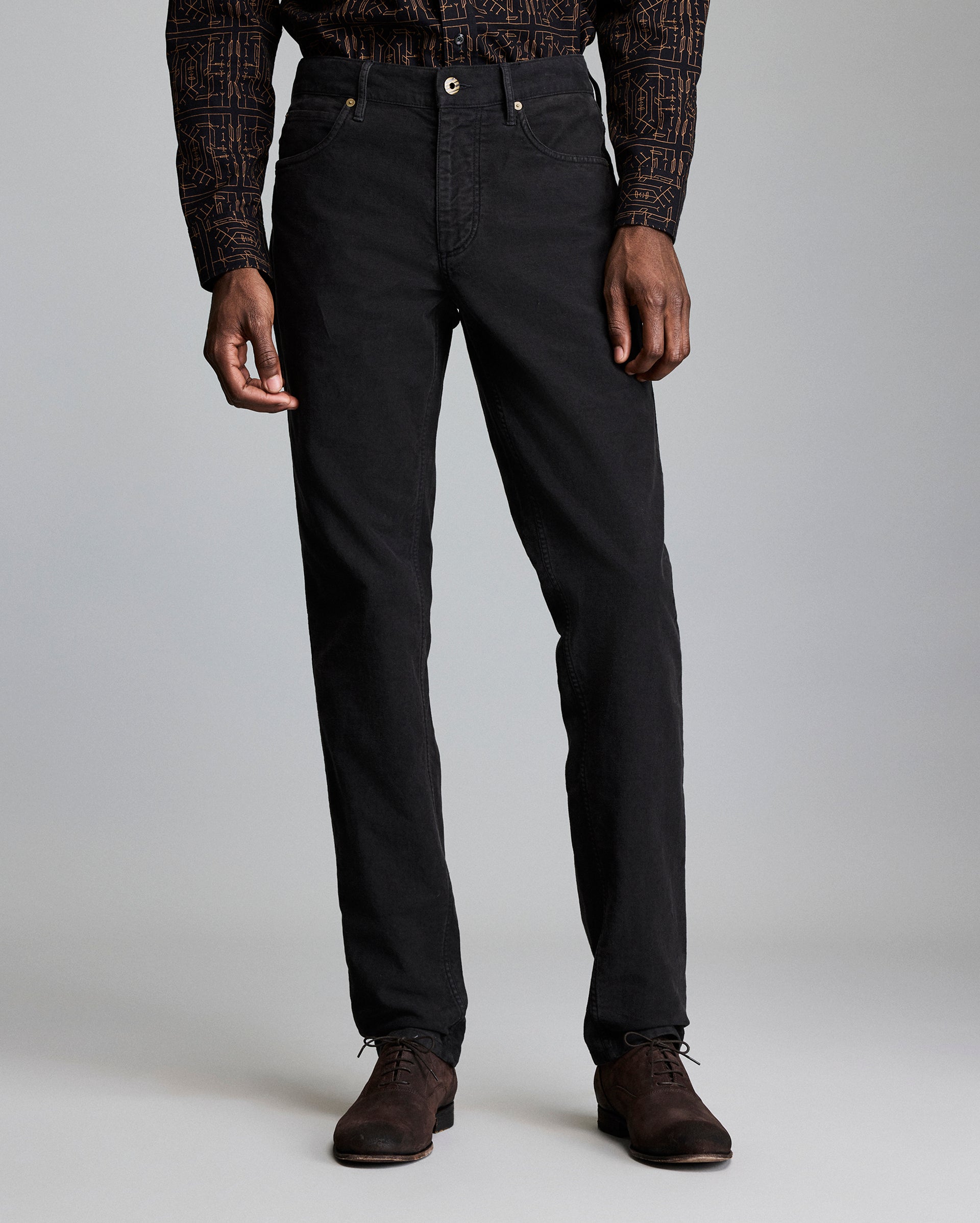 A man stands against a plain background wearing black pants, a patterned long-sleeve shirt, and dark brown dress shoes. His arms are relaxed at his sides, and his head is not visible in the frame.