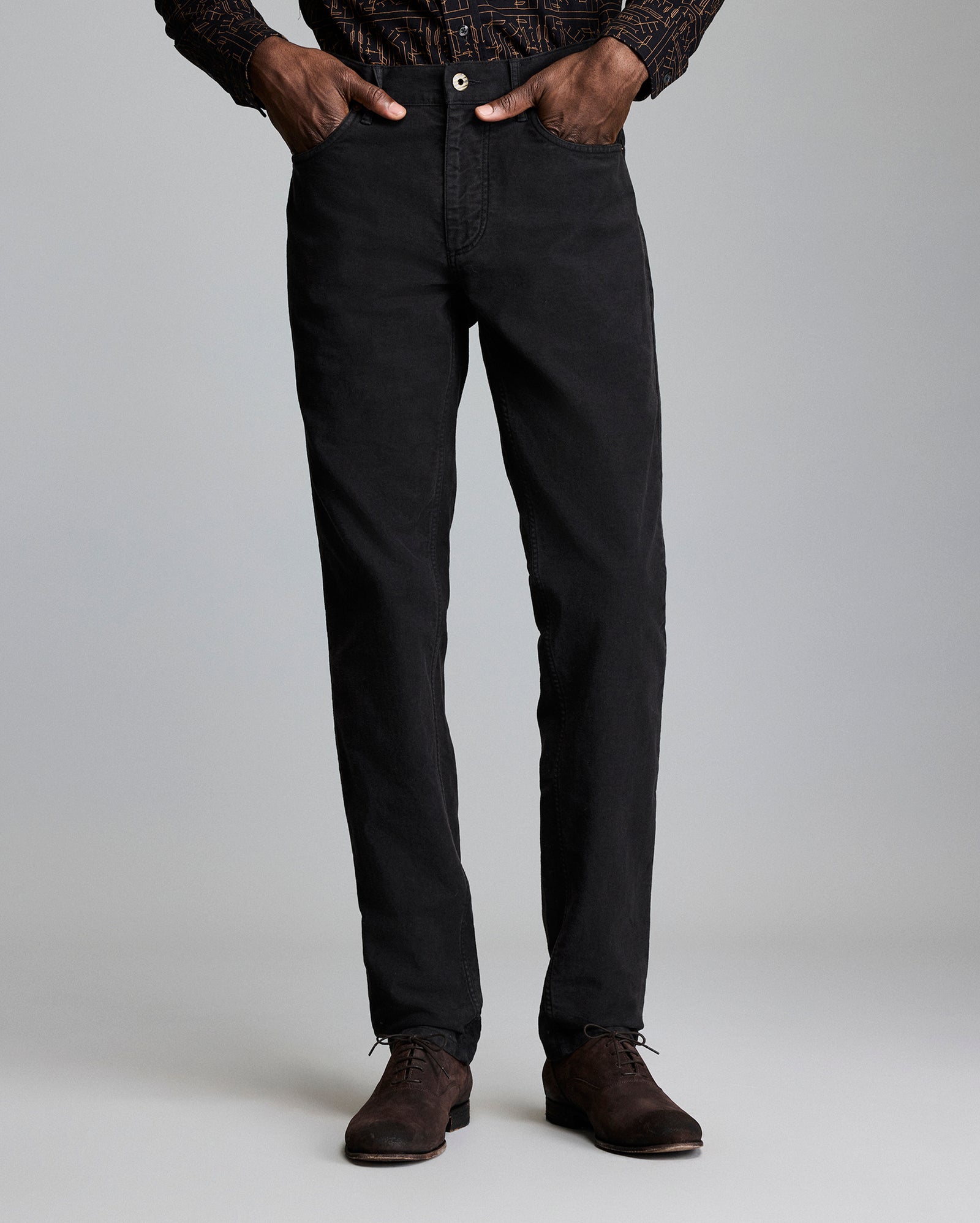A person stands against a plain background, wearing black straight-leg pants, a patterned long-sleeve shirt, and brown dress shoes. Their hands are in their pockets, and their upper body is partially out of frame.