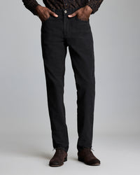 A person stands against a plain background, wearing black straight-leg pants, a patterned long-sleeve shirt, and brown dress shoes. Their hands are in their pockets, and their upper body is partially out of frame.