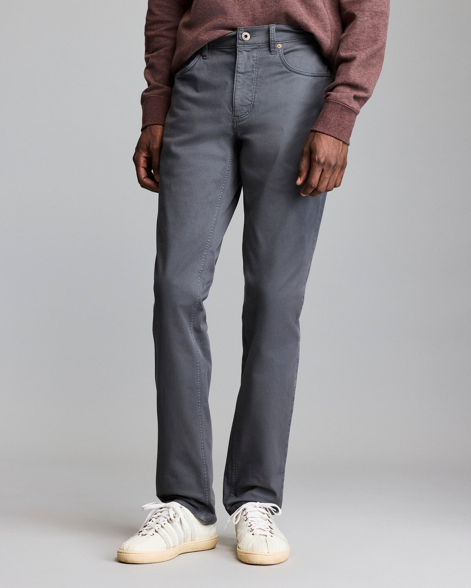 A person wearing a maroon sweater, gray pants, and white sneakers stands against a plain gray background. Only the lower half of their body is visible.