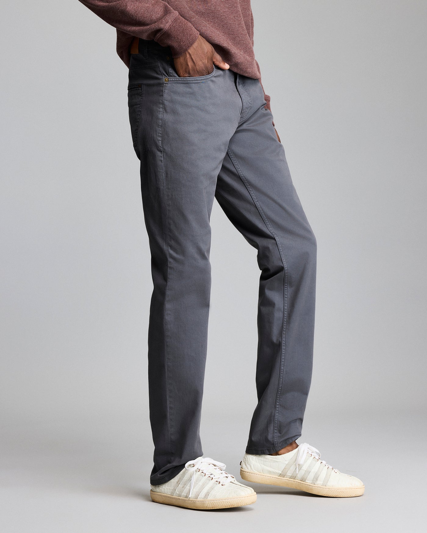 A person wearing a maroon long-sleeve shirt, gray pants, and white sneakers stands with one hand in their pocket against a plain gray background. The image shows the person from the waist down.