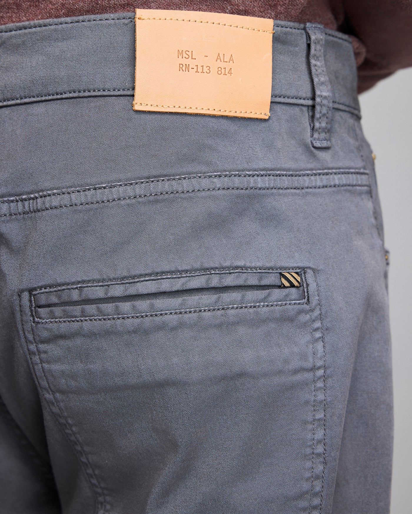 Close-up of the back of gray pants with a brown leather patch on the waistband displaying text MSL - ALA RN-113 814 and the top part of a back pocket with a small striped tag.