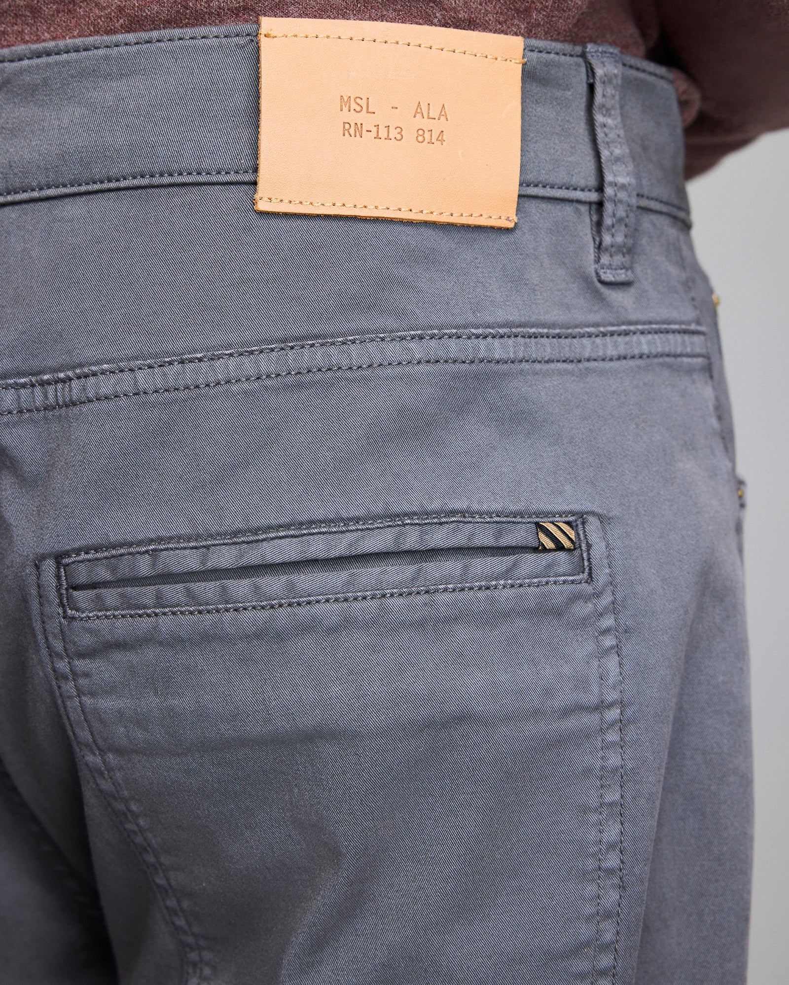 Close-up of the back of gray pants with a brown leather patch on the waistband displaying text MSL - ALA RN-113 814 and the top part of a back pocket with a small striped tag.