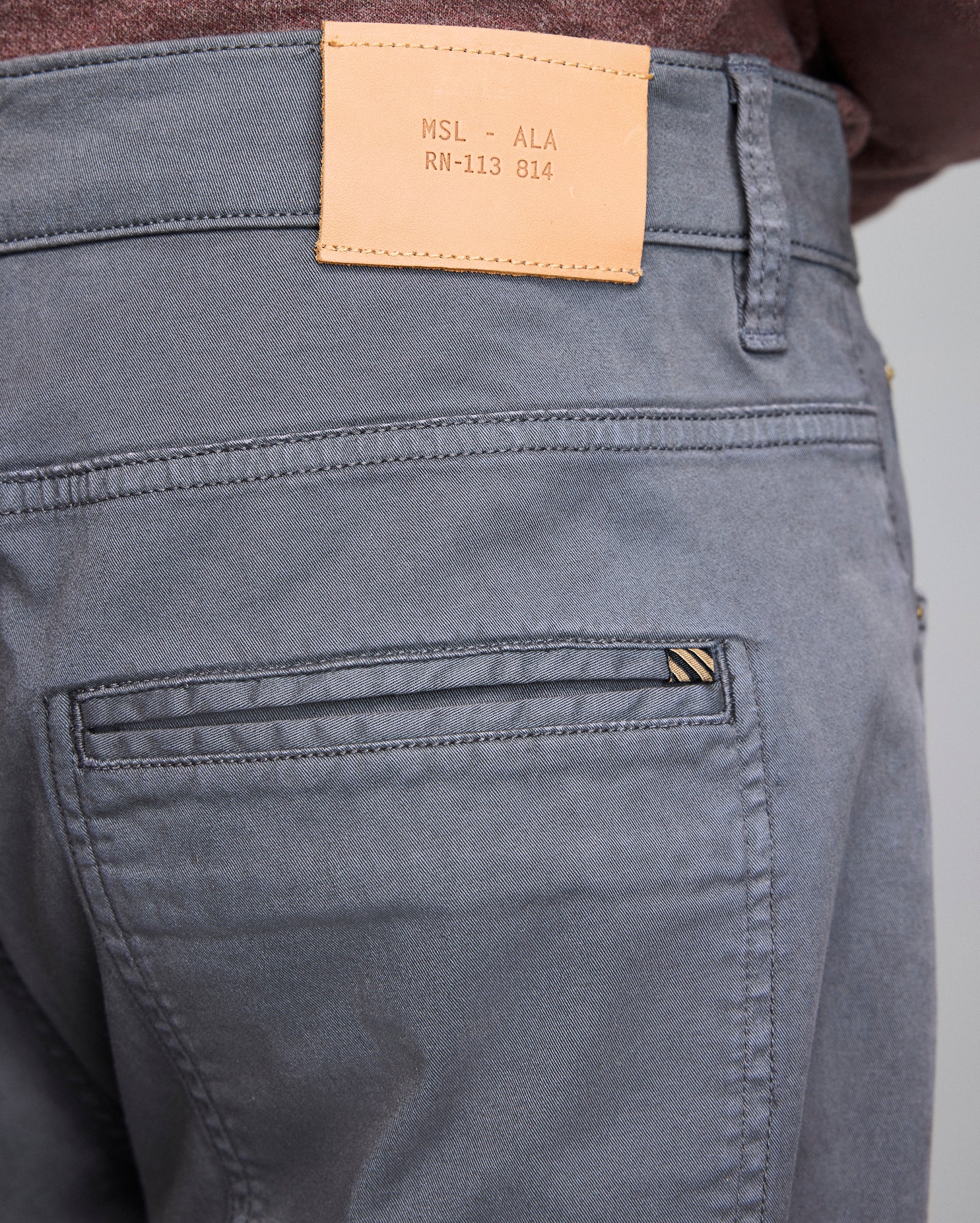 Close-up of the back of gray pants with a brown leather patch on the waistband displaying text MSL - ALA RN-113 814 and the top part of a back pocket with a small striped tag.