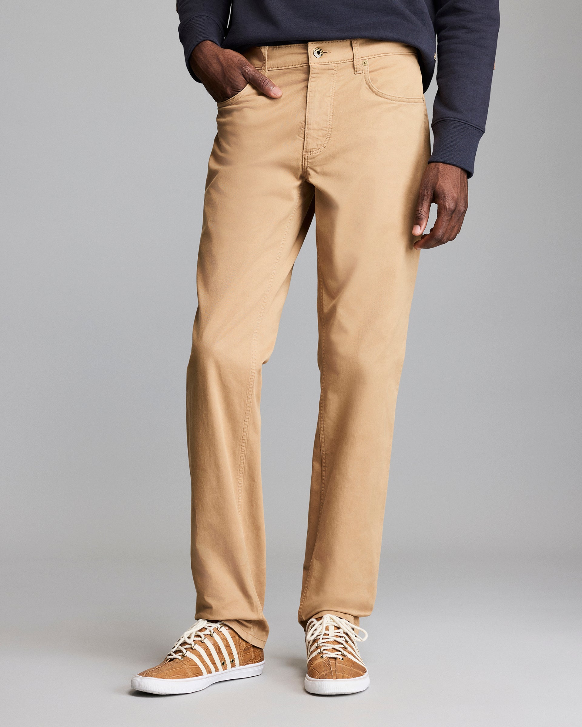 A person wearing tan straight-leg pants, a dark long-sleeve shirt, and tan sneakers with white laces stands against a plain gray background. Only the lower half of the body is visible.