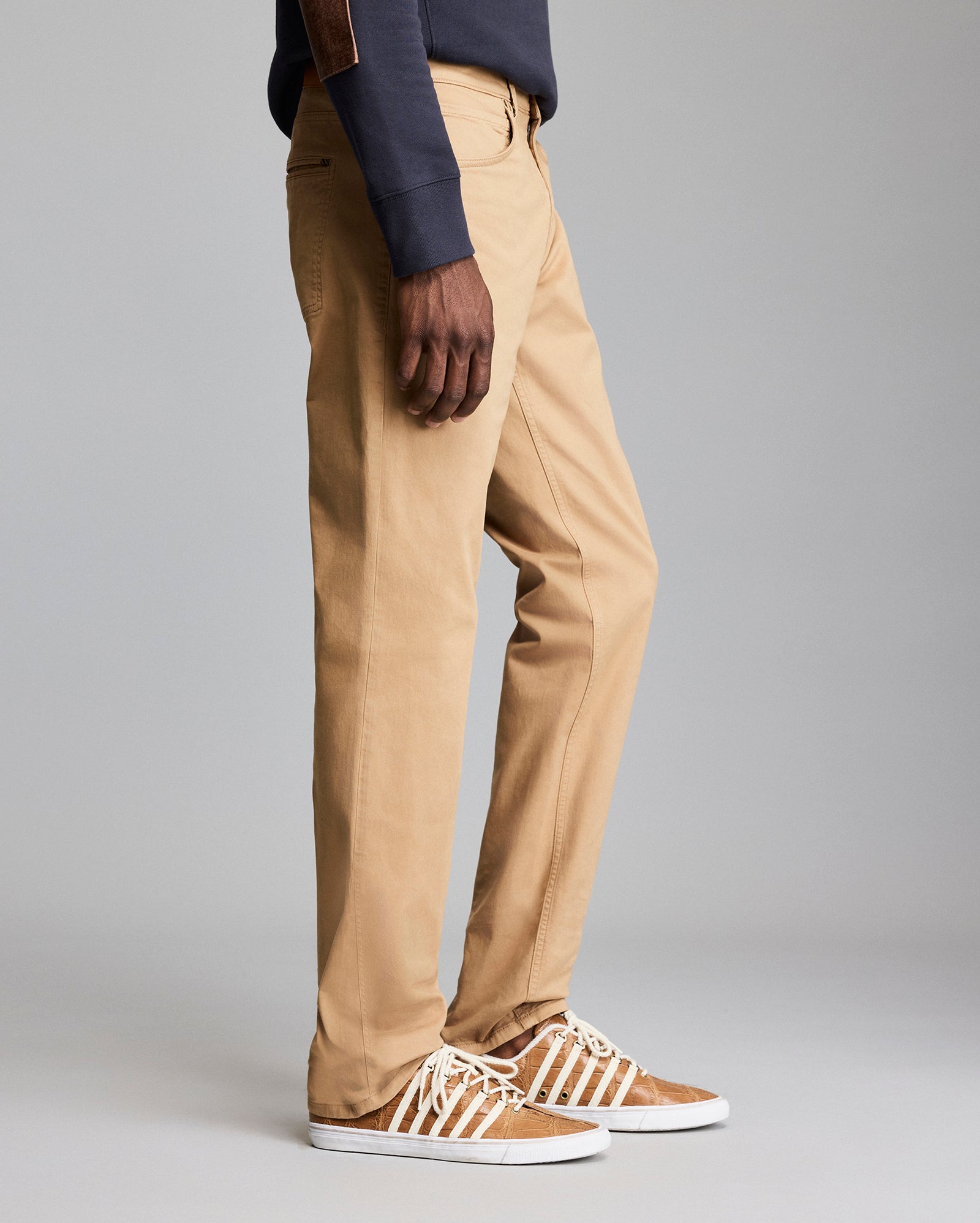 A person wearing tan khaki pants, a dark long-sleeve shirt, and brown sneakers with white laces is standing against a plain gray background. Only the lower half of the body is visible.