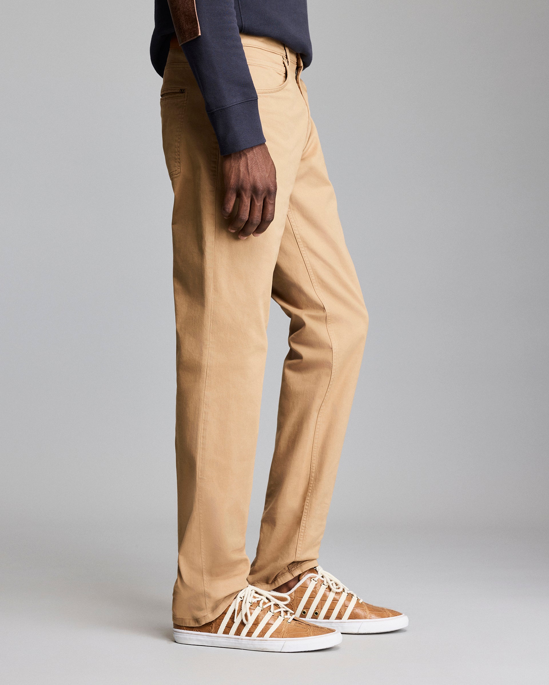 A person wearing tan khaki pants, a dark long-sleeve shirt, and brown sneakers with white laces is standing against a plain gray background. Only the lower half of the body is visible.