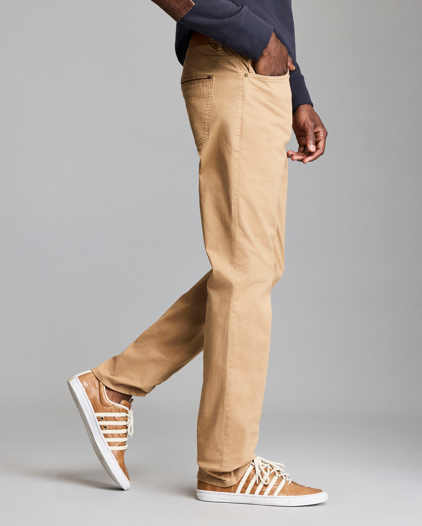 A person wearing beige chinos, a navy sweatshirt, and tan sneakers with white laces, shown in a side view standing against a plain gray background.