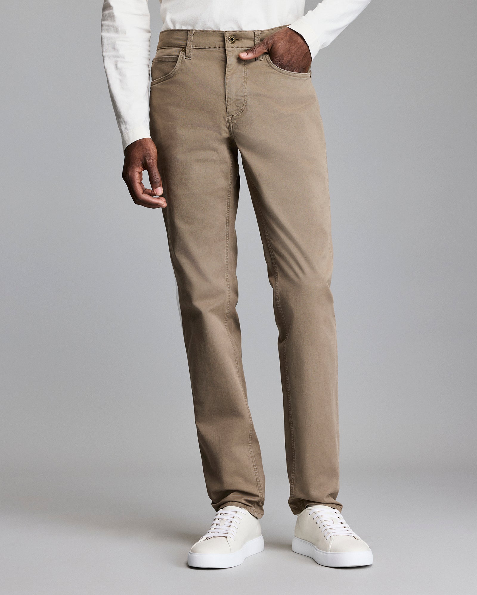 A person wearing light brown pants, a white long-sleeve shirt, and white sneakers stands against a plain gray background. The photo is cropped from the shoulders down.