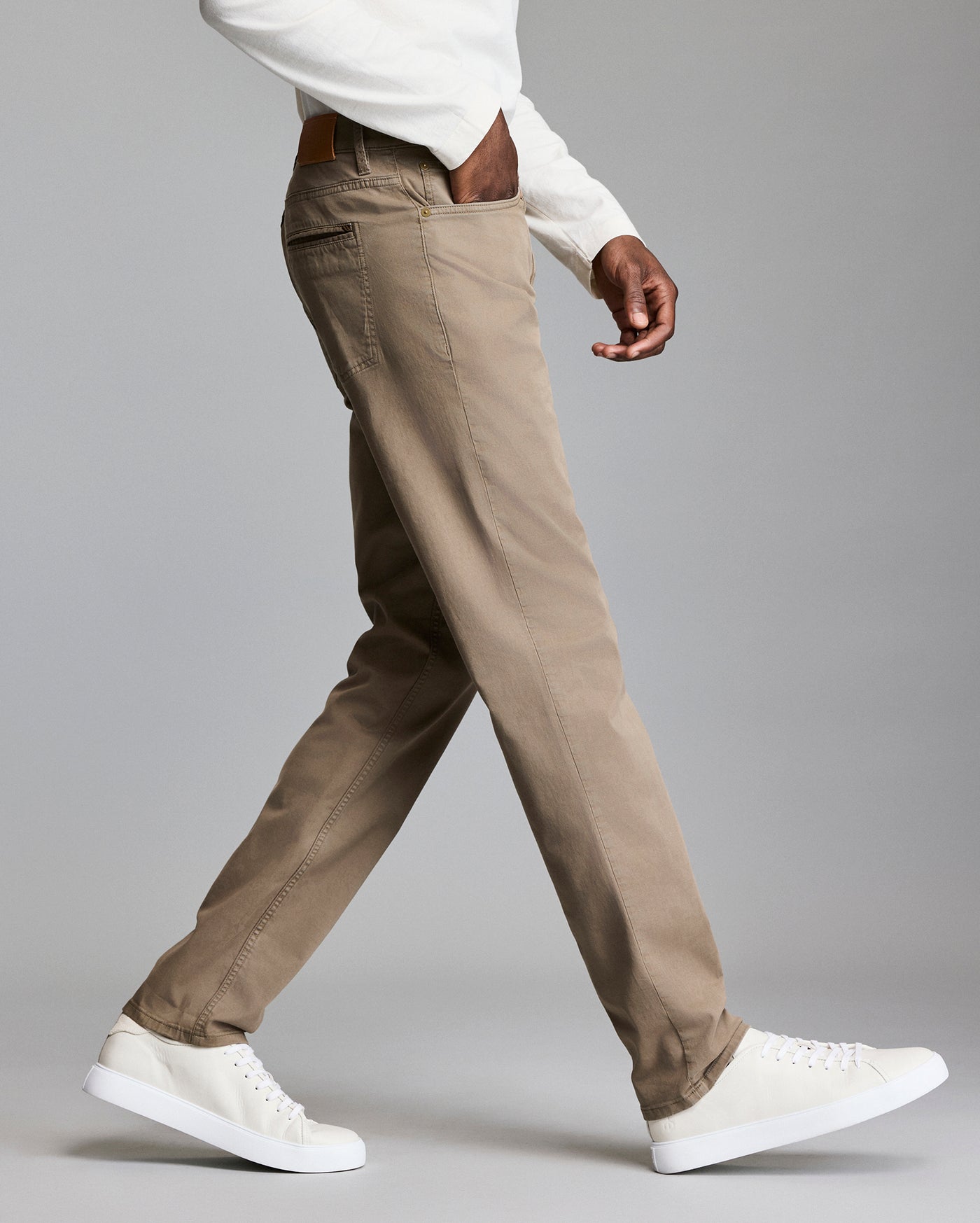A person wearing light brown pants, a white long-sleeve shirt, and white sneakers is walking against a plain gray background. Only the lower half of their body is visible.