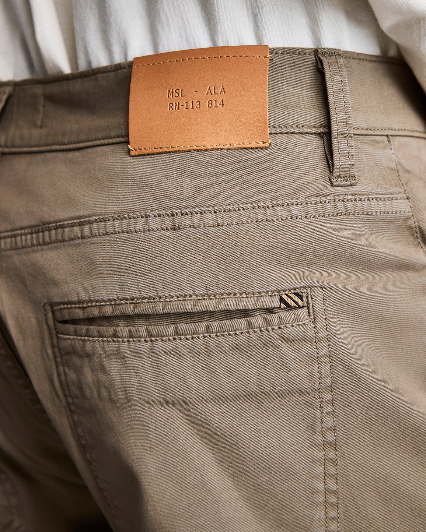 Close-up of the back waistband and pocket of khaki pants. A tan leather patch with the text MSL • ALA RN-113 814 is sewn above the pocket. A white shirt is partially visible above the pants.