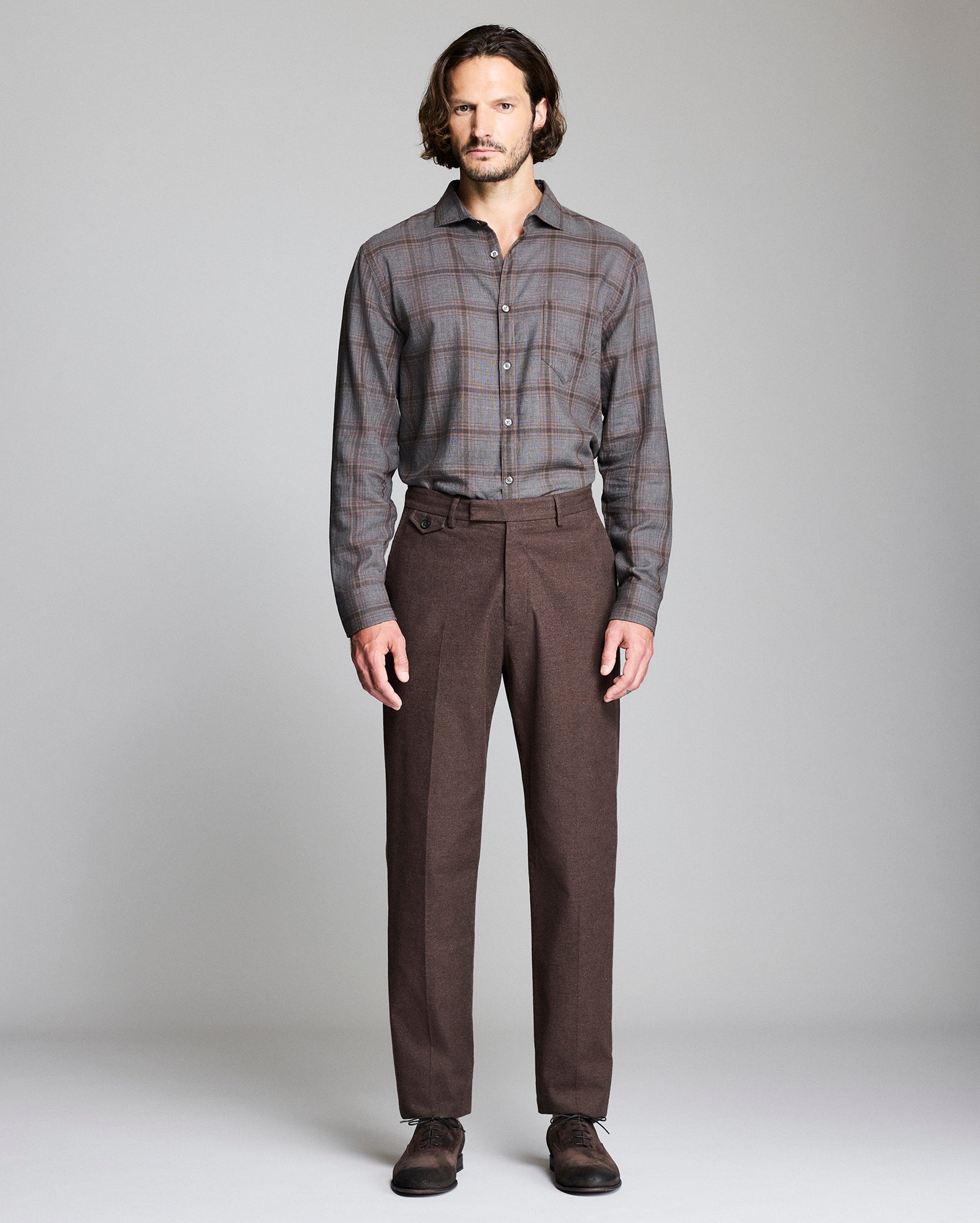 A man stands facing forward against a plain gray background, wearing a gray plaid button-up shirt tucked into brown dress pants, with dark brown shoes.