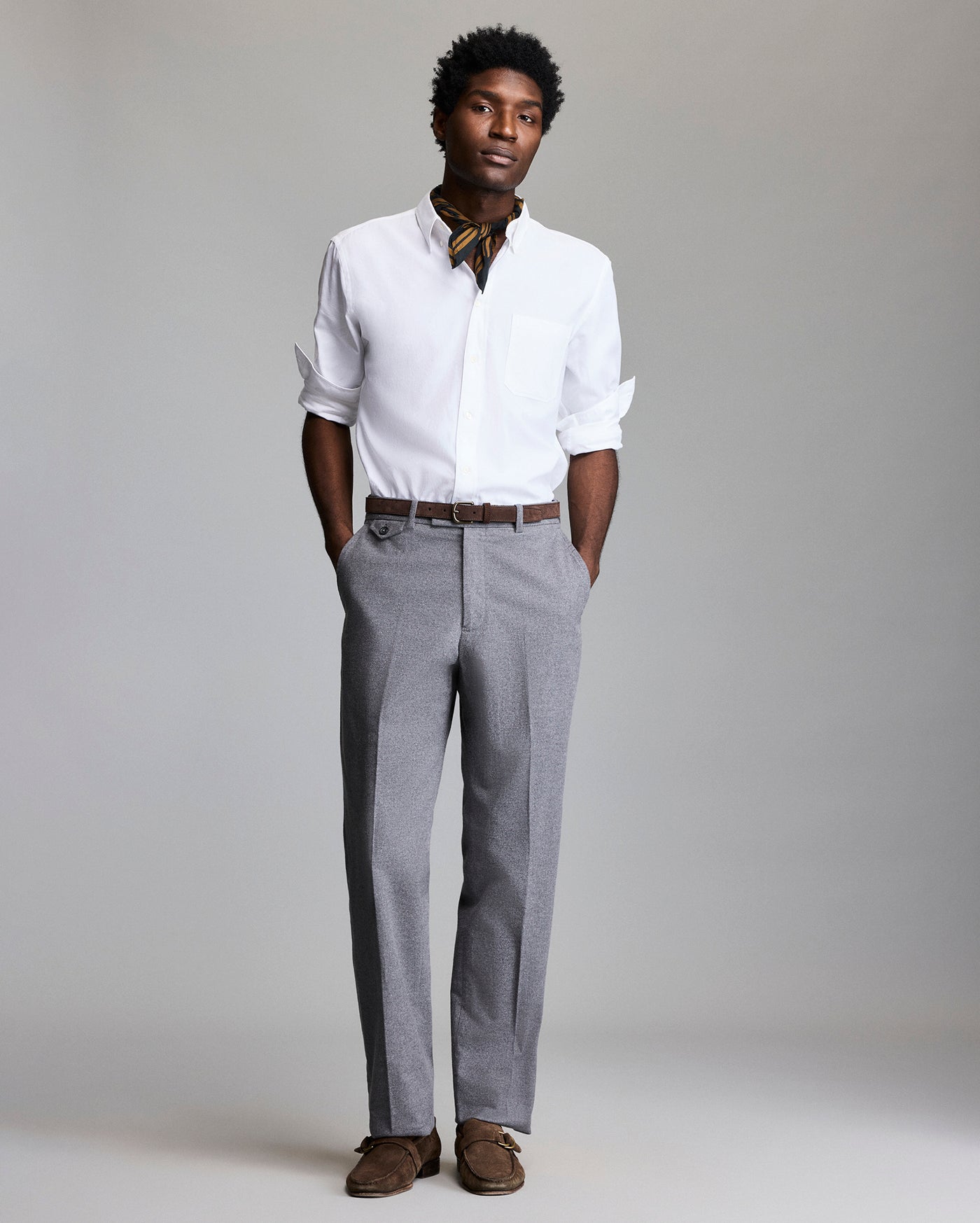 A man stands with hands in his pockets, wearing a white dress shirt with rolled-up sleeves, gray dress pants, a brown belt, brown shoes, and a patterned neck scarf, against a plain gray background.