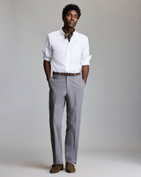 A man stands with hands in his pockets, wearing a white dress shirt with rolled-up sleeves, gray dress pants, a brown belt, brown shoes, and a patterned neck scarf, against a plain gray background.