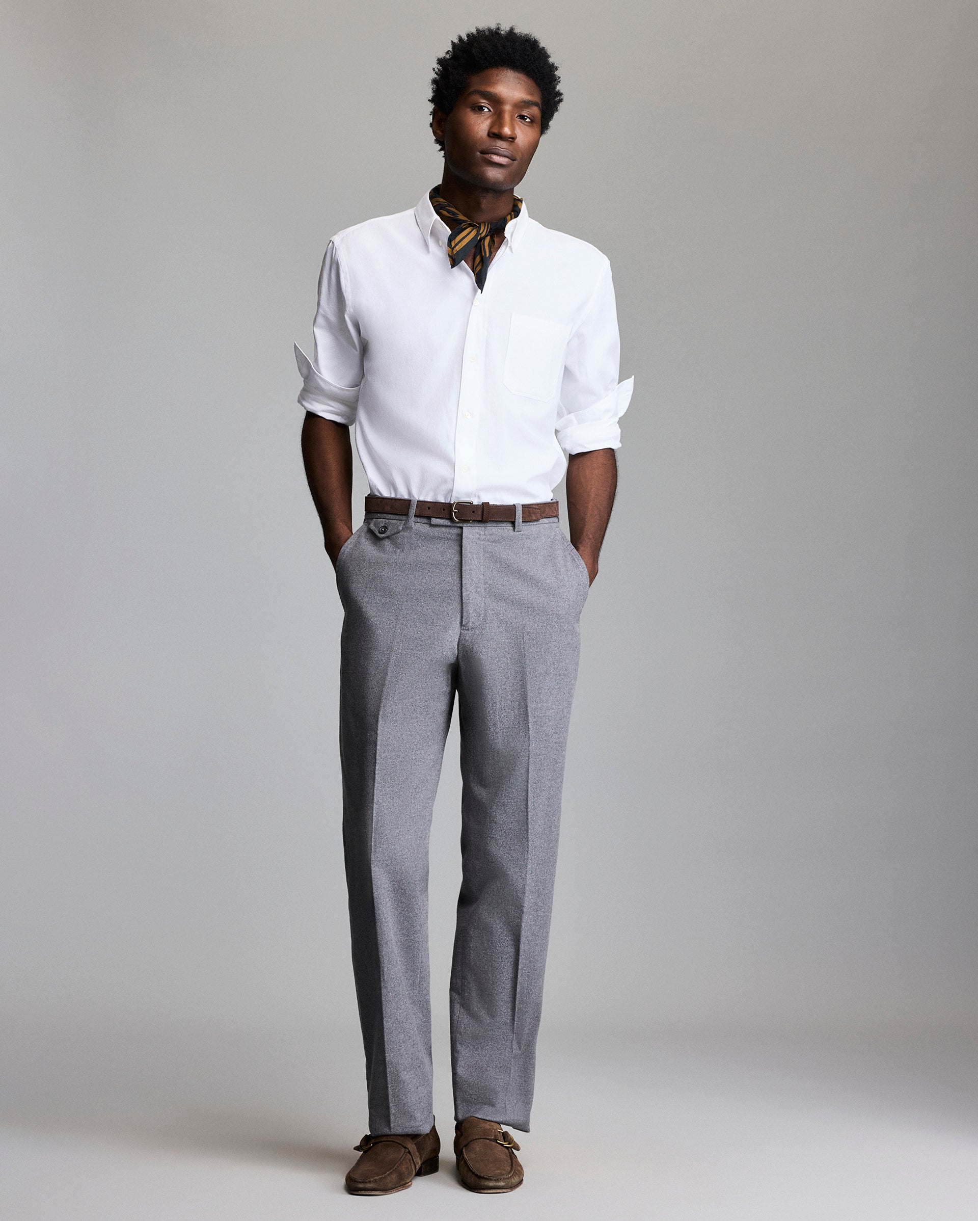 A man stands with hands in his pockets, wearing a white dress shirt with rolled-up sleeves, gray dress pants, a brown belt, brown shoes, and a patterned neck scarf, against a plain gray background.