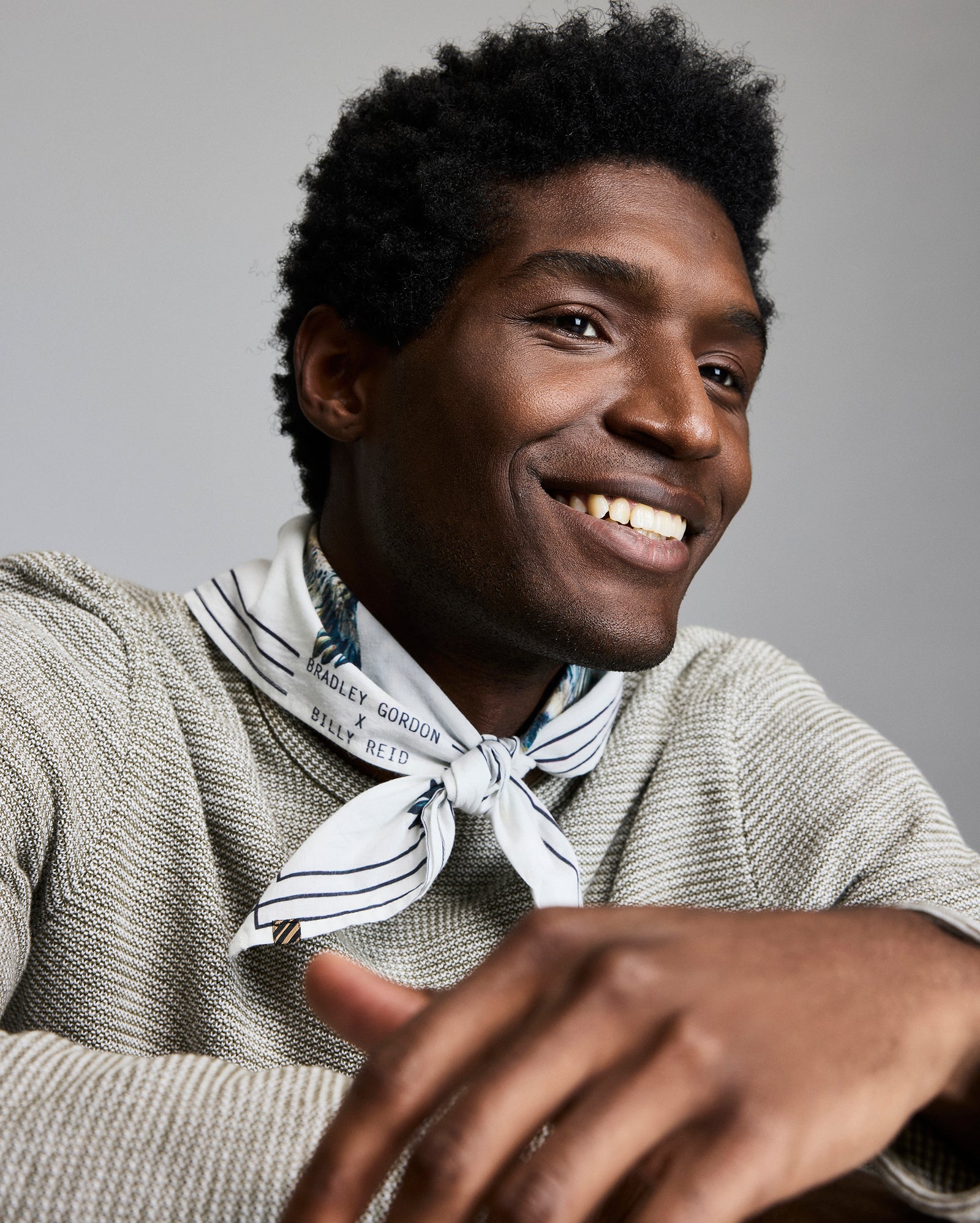 A smiling man with short curly hair wears a textured light sweater and a white striped scarf around his neck. He is looking off to the side, with his arm resting casually in front of him. The background is plain and neutral.