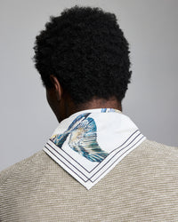 A person with short curly black hair, seen from behind, wears a textured beige jacket and a white scarf with blue and green fish print and dark stripes around the edges.