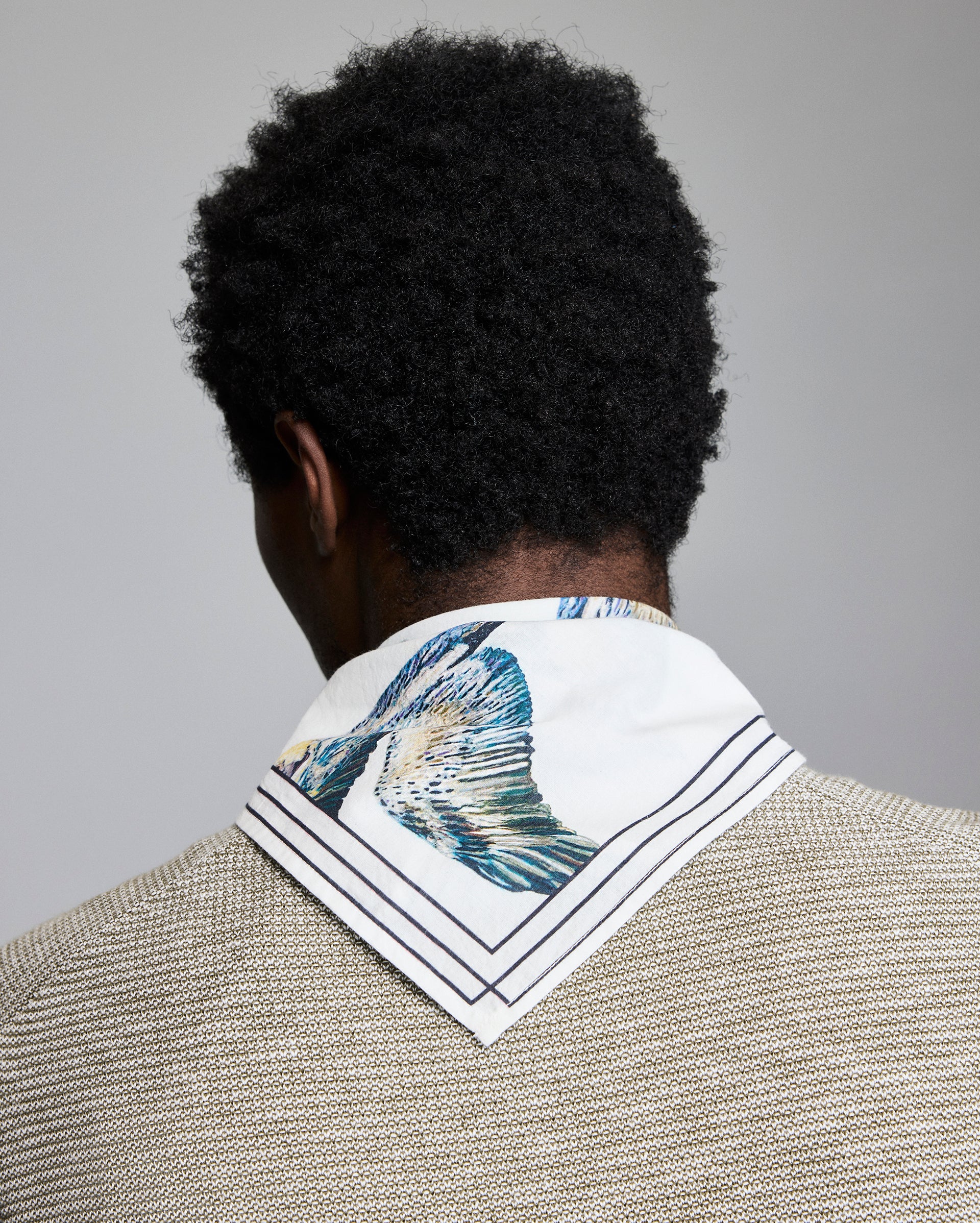 A person with short curly black hair, seen from behind, wears a textured beige jacket and a white scarf with blue and green fish print and dark stripes around the edges.