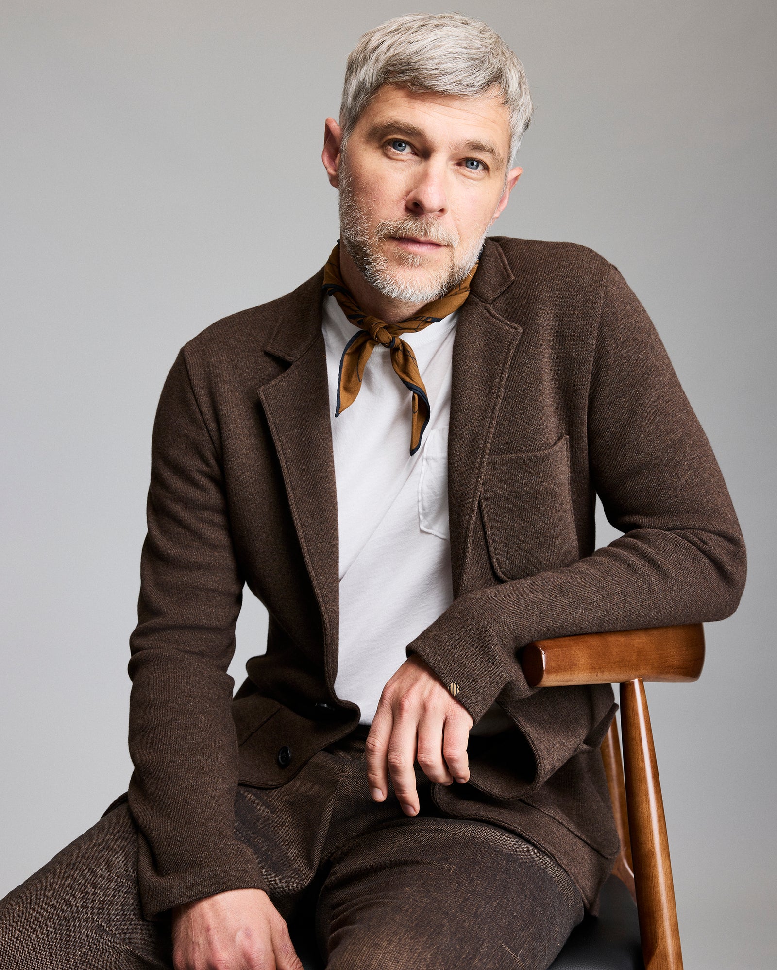 A man with short gray hair and a beard sits on a wooden chair, wearing a brown suit, white shirt, and a brown scarf tied around his neck, looking calmly at the camera against a plain gray background.