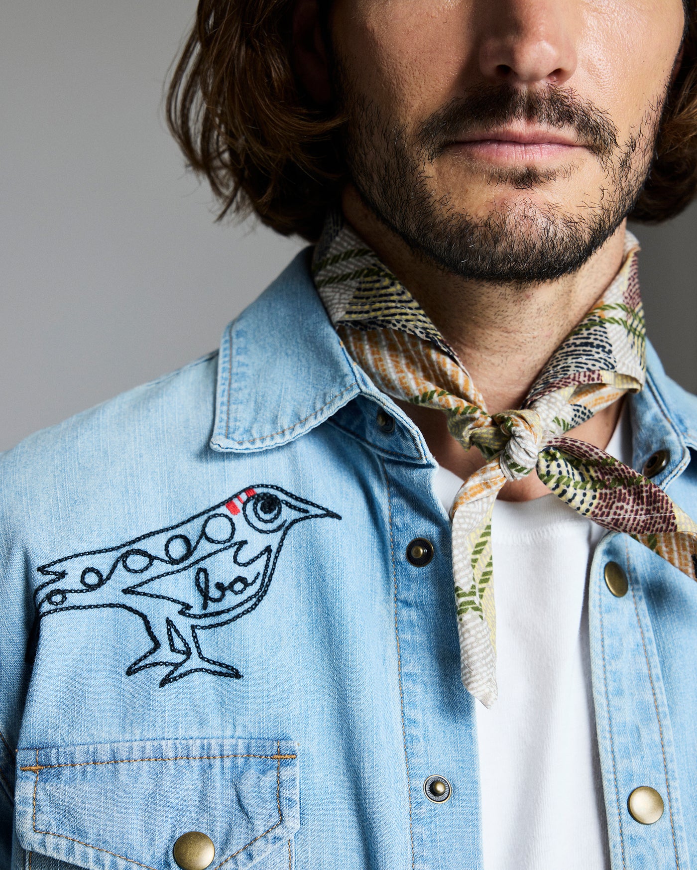 A man with medium-length brown hair and a trimmed beard wears a light denim shirt embroidered with a black bird and a patterned scarf tied around his neck. Only the lower half of his face and upper torso are visible.