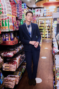 A person in a navy blue suit and checked shirt stands in the aisle of a brightly lit convenience store, surrounded by colorful cereal boxes and snack products on the shelves.