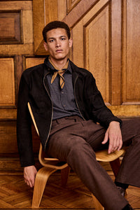 A man sits on a wooden chair in a room with wooden paneled walls. He wears brown pants, a checked shirt, a black jacket, and a light brown neck scarf, looking confidently at the camera.