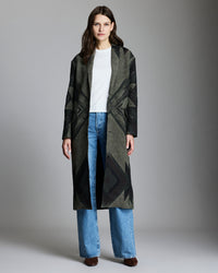 A woman stands facing forward, wearing a long dark patterned coat over a white top and blue jeans. She has long brown hair and is wearing brown shoes, standing against a plain gray background.
