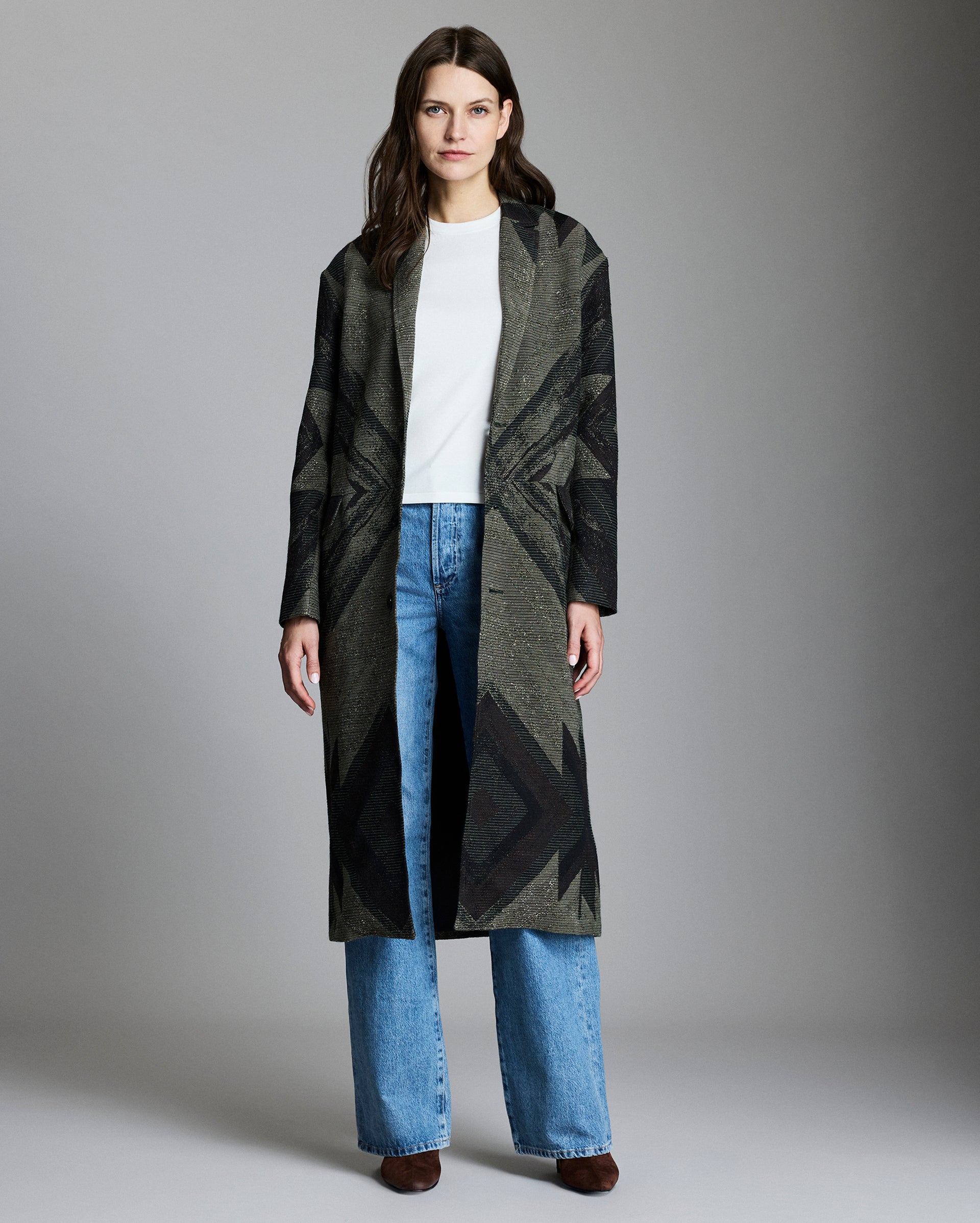 A woman stands facing forward, wearing a long dark patterned coat over a white top and blue jeans. She has long brown hair and is wearing brown shoes, standing against a plain gray background.
