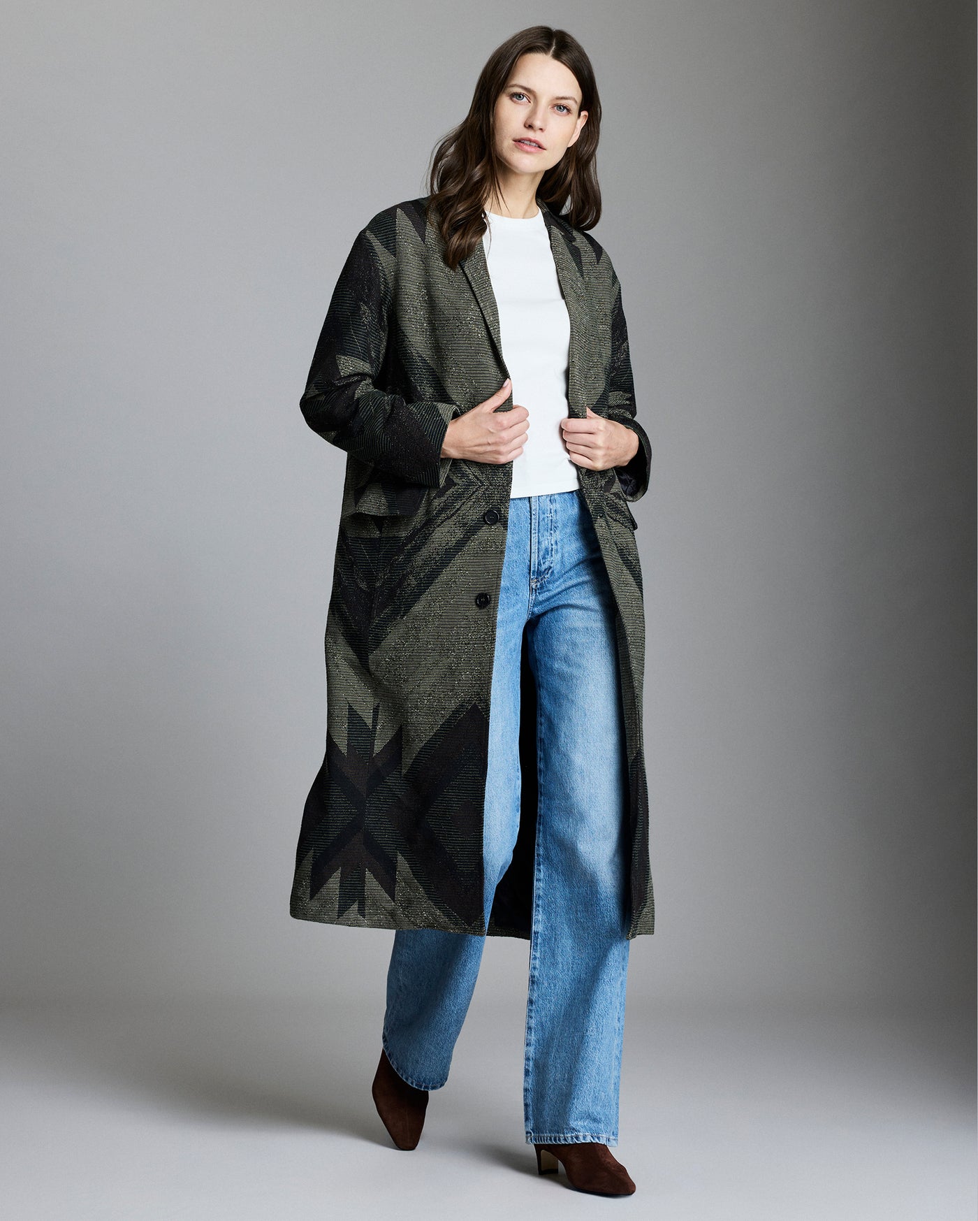 Woman in a patterned long coat, white top, blue jeans, and brown shoes stands against a plain gray background, looking forward with a neutral expression.