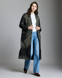 Woman in a patterned long coat, white top, blue jeans, and brown shoes stands against a plain gray background, looking forward with a neutral expression.