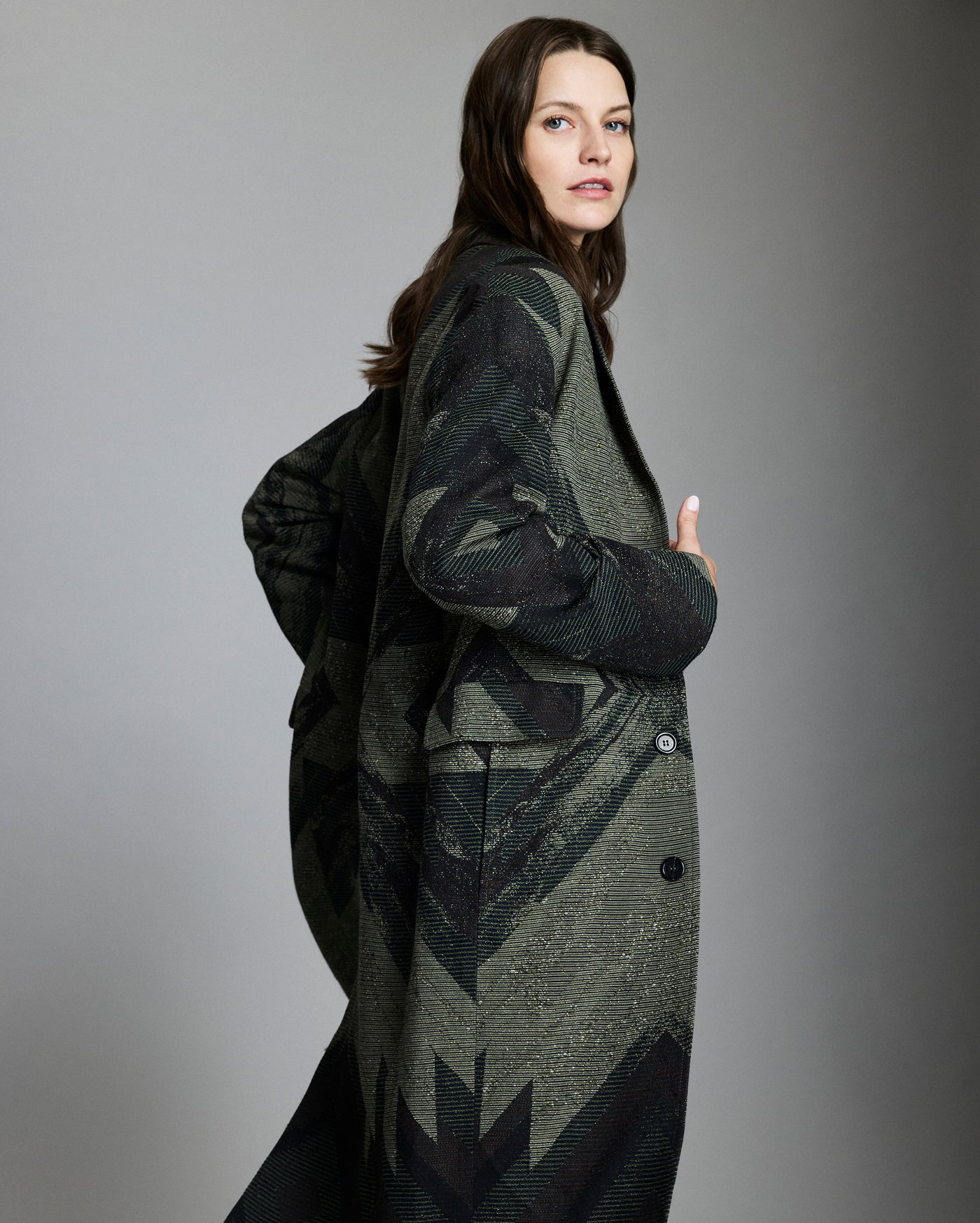 A woman with long brown hair wears a dark, patterned overcoat, standing sideways and looking at the camera against a plain, light gray background.