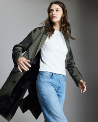 A woman with long brown hair wears a patterned green coat over a white shirt and blue jeans, standing against a gray background with one arm raised and looking to the side.