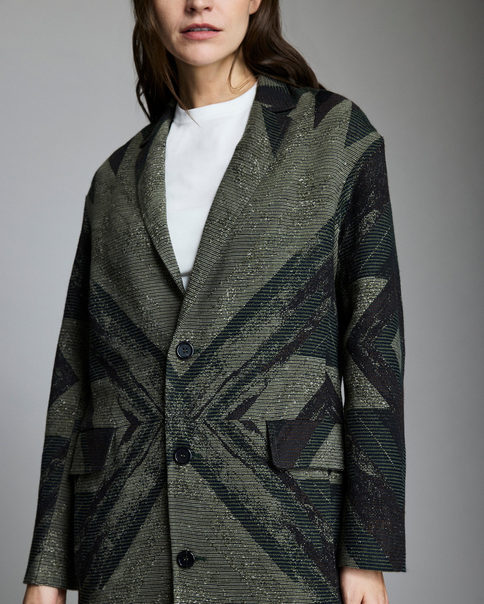 A woman wearing a patterned, button-up coat with black and olive green geometric designs over a white top, shown from mid-thigh up against a plain gray background.