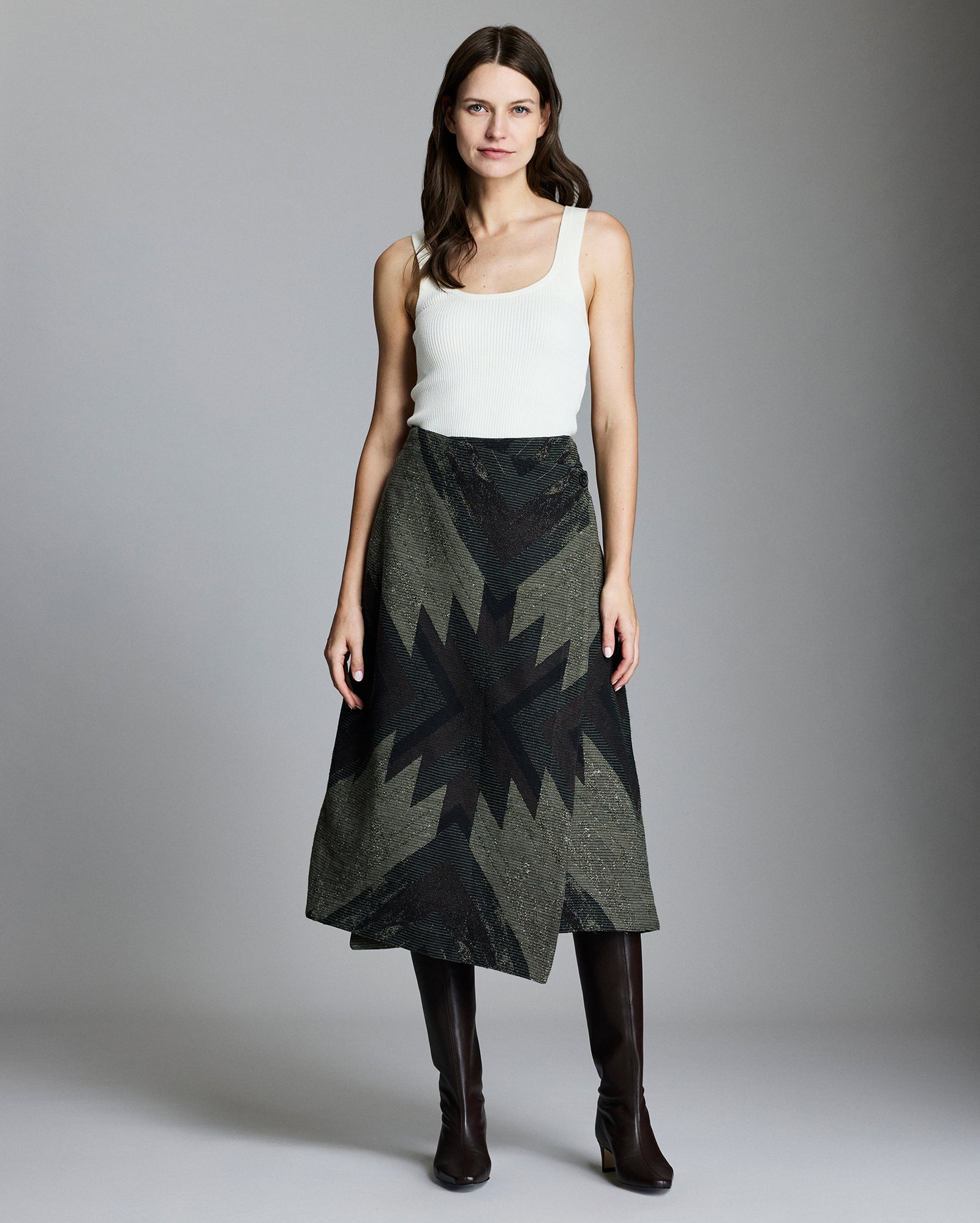 A woman stands against a plain background, wearing a white sleeveless top, a dark patterned A-line skirt with a geometric design, and knee-high black boots. She has long brown hair and a neutral expression.