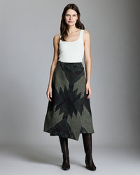 A woman stands against a plain background, wearing a white sleeveless top, a dark patterned A-line skirt with a geometric design, and knee-high black boots. She has long brown hair and a neutral expression.