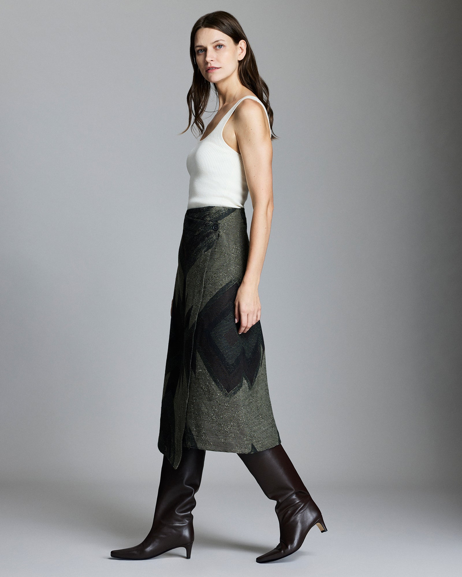 A woman with long brown hair wears a white sleeveless top, an olive green wrap skirt with a dark geometric pattern, and tall brown leather boots, standing in profile against a plain gray background.