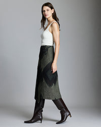 A woman with long brown hair wears a white sleeveless top, an olive green wrap skirt with a dark geometric pattern, and tall brown leather boots, standing in profile against a plain gray background.