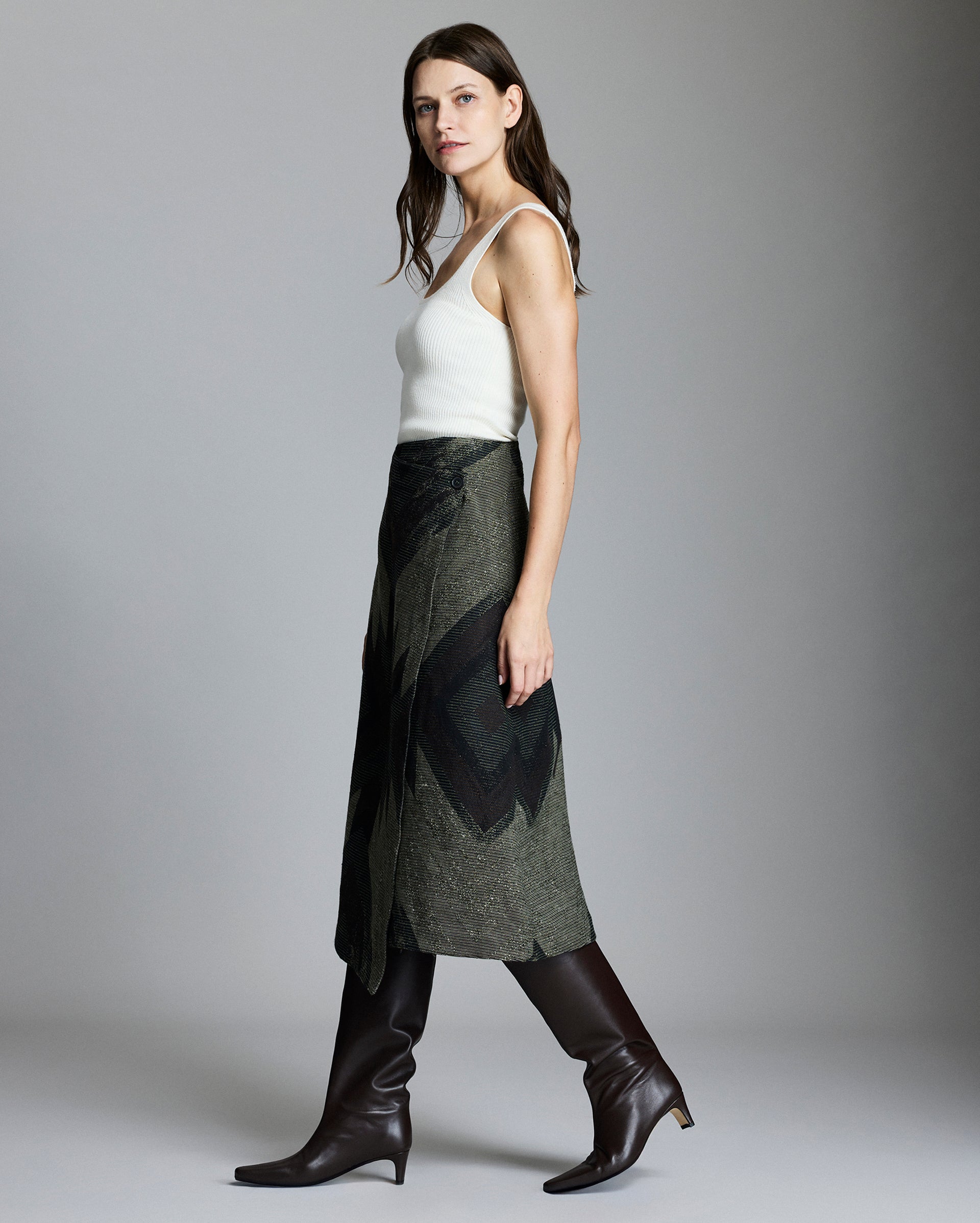 A woman with long brown hair wears a white sleeveless top, an olive green wrap skirt with a dark geometric pattern, and tall brown leather boots, standing in profile against a plain gray background.