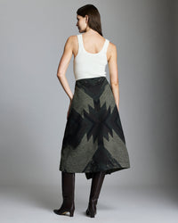 A woman stands facing away, wearing a white sleeveless top, an olive green skirt with a black geometric star pattern, and knee-high dark brown boots. The background is plain and gray.