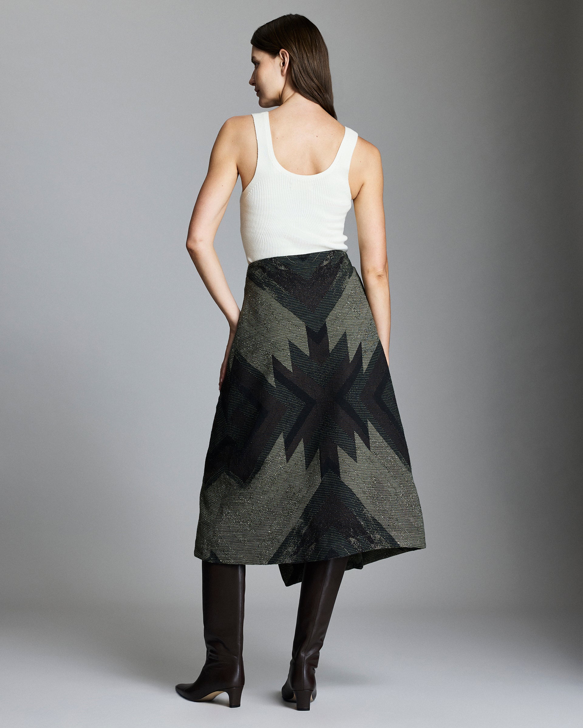 A woman stands facing away, wearing a white sleeveless top, an olive green skirt with a black geometric star pattern, and knee-high dark brown boots. The background is plain and gray.