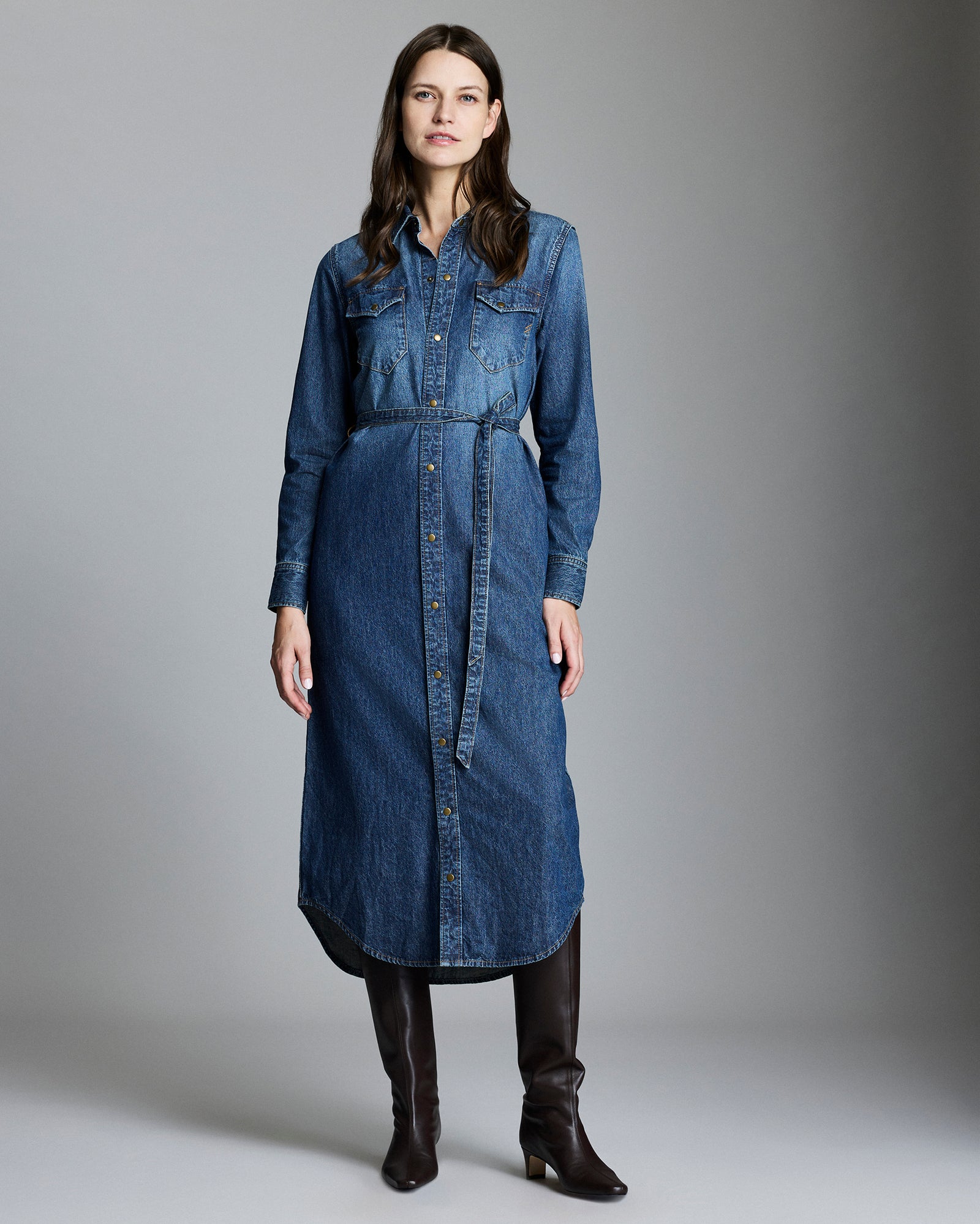 A woman stands against a plain gray background wearing a long-sleeve, button-down denim dress with a belt, and knee-high brown boots. She has long, wavy brown hair and a neutral expression.