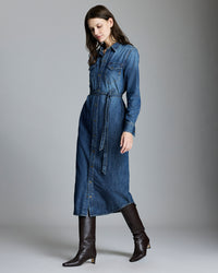 A woman wearing a long-sleeved, button-down denim dress with a waist tie and dark knee-high boots stands against a plain gray background, looking down and slightly to the side.