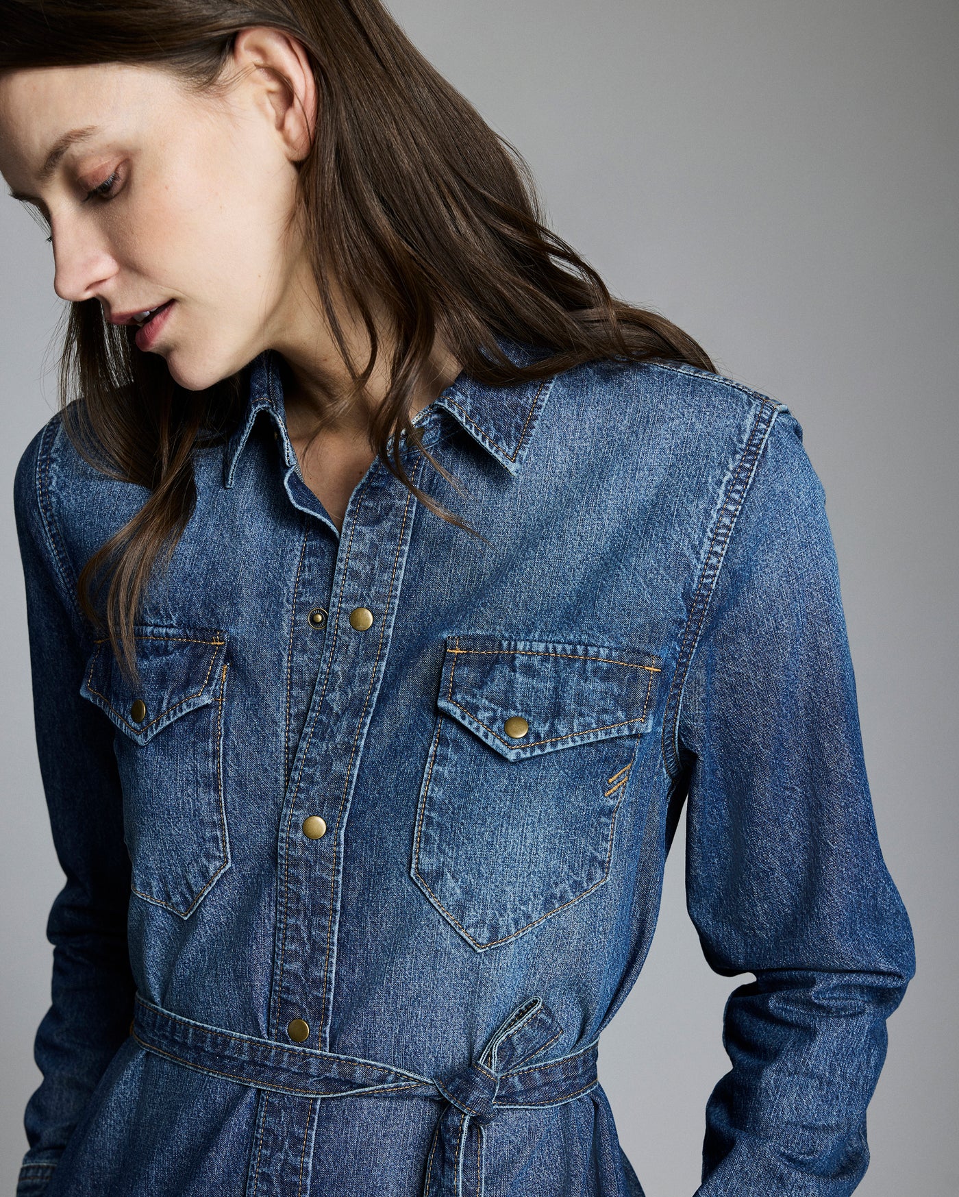 A woman with long brown hair wearing a blue denim button-up shirt dress with front pockets and a tied belt, looking down against a plain grey background.