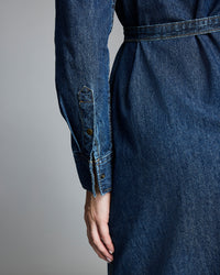 A close-up of a person’s hand resting at their side, wearing a long-sleeved, belted denim dress, shown from the back with details of the sleeve cuff and stitching visible.
