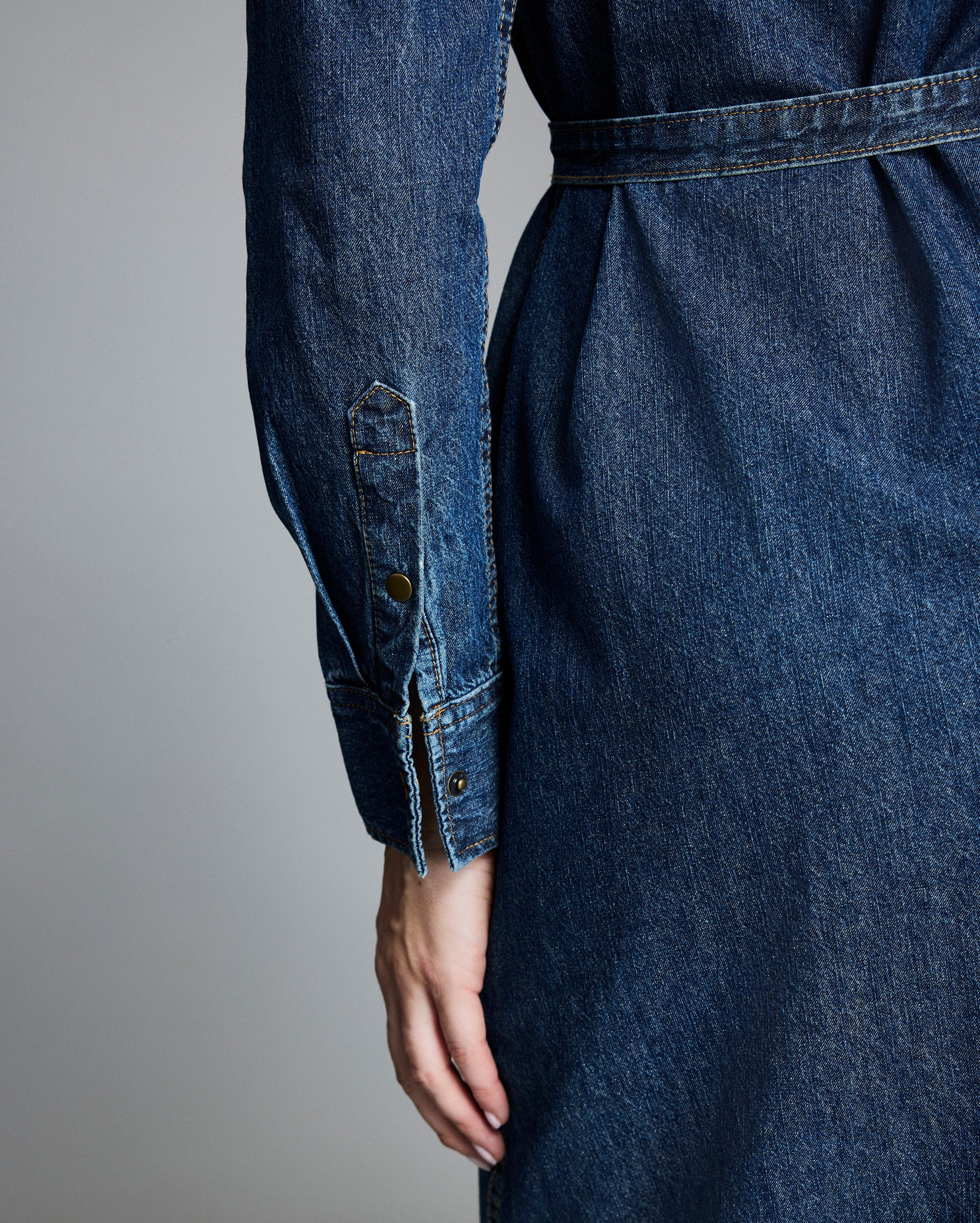 A close-up of a person’s hand resting at their side, wearing a long-sleeved, belted denim dress, shown from the back with details of the sleeve cuff and stitching visible.