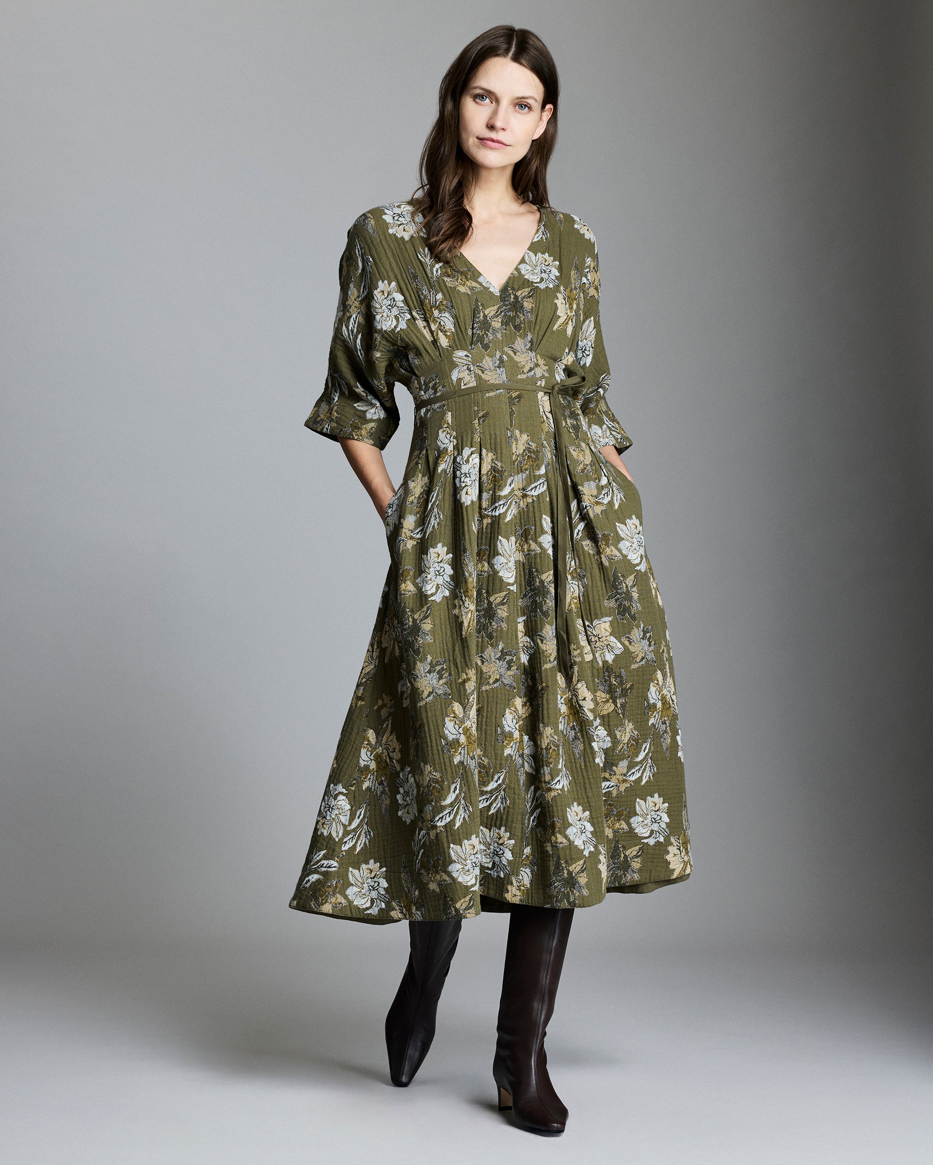 A woman stands against a gray background wearing an olive green, floral midi dress with a V-neckline and three-quarter sleeves, paired with knee-high dark brown boots. She has long brown hair and her hands in her pockets.